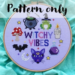 May include: A cross-stitch pattern in a wooden hoop, featuring a witchy theme. The design includes a cauldron, frog, mushroom, bat, cat, and the words "Witchy Vibes." The background is a light purple colour, with stars and a crescent moon.
