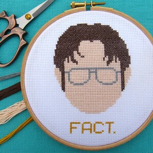 May include: A brown and white cross-stitch embroidery of a person with brown hair wearing glasses. The embroidery is framed in a natural wood hoop and the text "FACT." is stitched in yellow below the image.