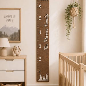 May include: A vertical wooden growth chart ruler featuring a dark brown finish with white numerical markings from 1 to 6 feet. The board includes white mountain and pine tree cutouts, and the text "The Harris Family" in white script.