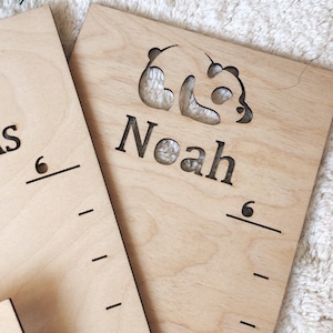 Pu&ograve; includere: Un grafico di crescita in legno con un ritaglio a forma di panda e il nome "Noah" inciso su di esso. Il grafico &egrave; contrassegnato per misurare l'altezza in centimetri.