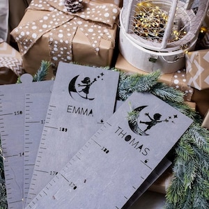 Pu&ograve; includere: Due grafici di crescita in legno grigio con un righello e una silhouette di un bambino che raggiunge una stella. I grafici sono personalizzati con i nomi "Emma" e "Thomas".