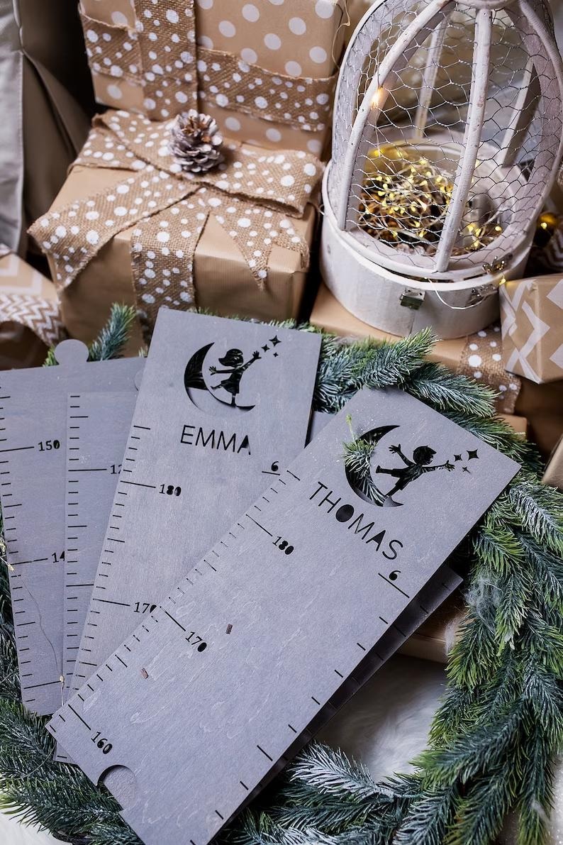 Pu&ograve; includere: Due grafici di crescita in legno grigio con un righello e una silhouette di un bambino che raggiunge una stella. I grafici sono personalizzati con i nomi "Emma" e "Thomas".
