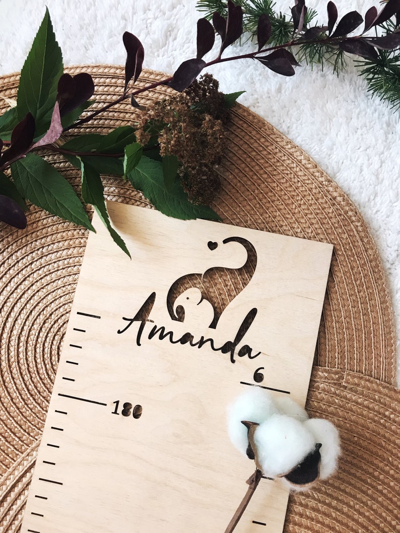 Pu&ograve; includere: Un grafico di crescita in legno con un ritaglio di un elefante e il nome "Amanda" inciso. Il grafico ha un righello sul lato con segni fino a 180 cm.