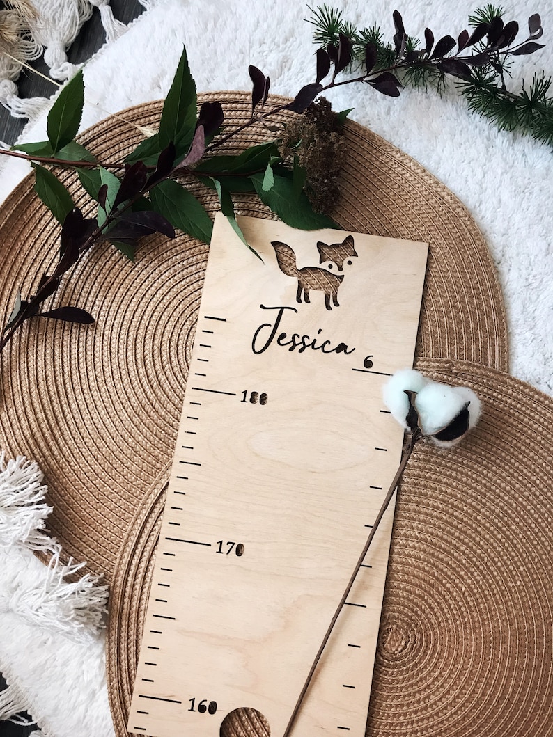 Pu&ograve; includere: Un grafico di crescita in legno con un ritaglio di volpe e il nome "Jessica" scritto su di esso. Il grafico ha segni per l'altezza in centimetri, a partire da 160 cm e fino a 180 cm.