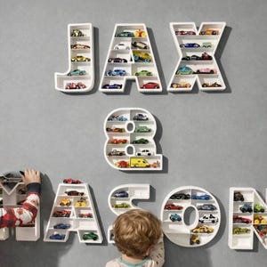 May include: White, letter-shaped shelves spelling "JAX" and "MASON", filled with toy cars in various colours. The number "8" is also a shelf. The shelves are mounted on a grey wall, creating a playful display.