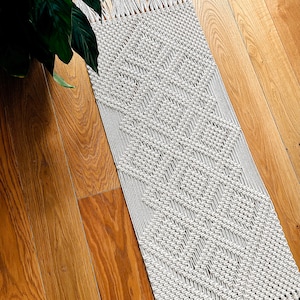 May include: A white macrame rug with a geometric diamond pattern and fringe on the ends. The rug is laid out on a wooden floor.