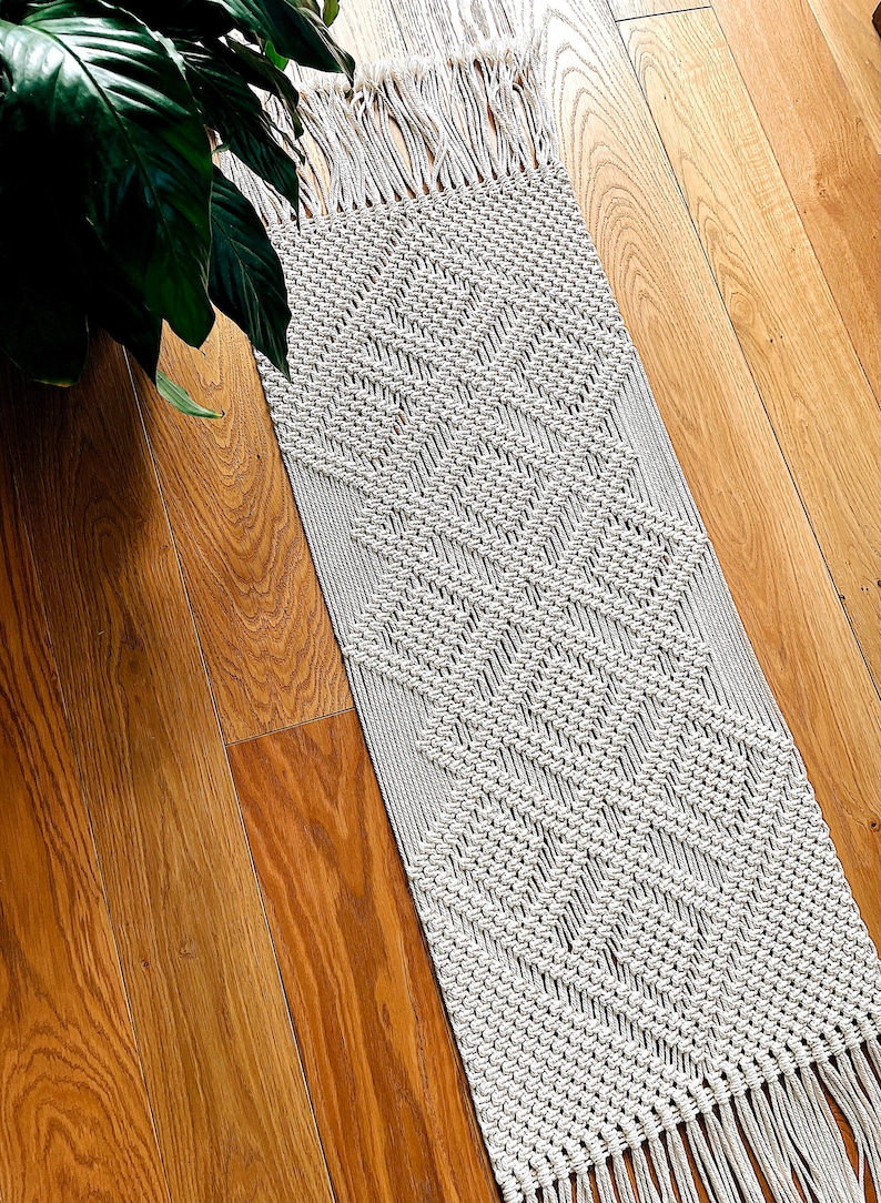 May include: A white macrame rug with a geometric diamond pattern and fringe on the ends. The rug is laid out on a wooden floor.