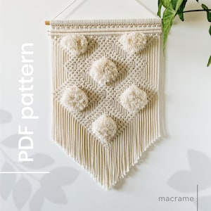 Könnte beinhalten: Ein beigefarbener Makramee-Wandbehang mit einem Rautenmuster und Pompons. Der Wandbehang ist aus natürlicher Baumwollschnur gefertigt und hat unten einen Fransen. Der Text "PDF pattern" ist vertikal auf der linken Seite des Bildes geschrieben. Der Text "macrame" ist horizontal am unteren Rand des Bildes geschrieben.