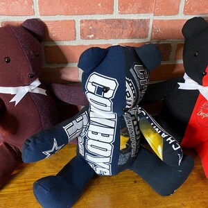 Puede incluir: Tres osos de peluche hechos a mano con camisetas recicladas. Los osos están sentados sobre una superficie de madera. Un oso es marrón, otro es azul marino con parches blancos y amarillos, y el último es negro y rojo.