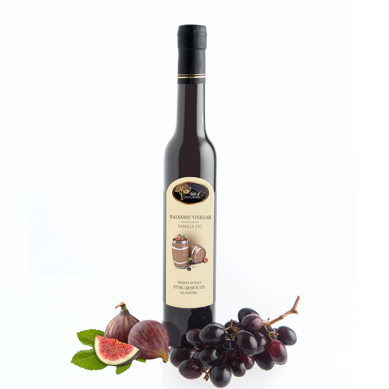 May include: A dark glass bottle of balsamic vinegar with a black cap and gold trim. The label reads "Balsamic Vinegar Infused with Vanilla Fig." Fresh figs and a bunch of grapes are arranged near the bottle.
