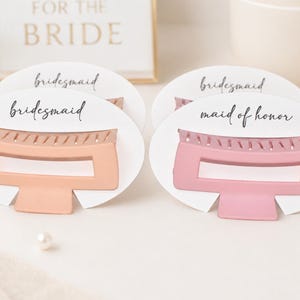 May include: Two hair claw clips, one peach and one pink, presented on white cards. The cards are labeled with the words "bridesmaid" and "maid of honor" in elegant script. The background includes a framed sign that says "For the Bride".