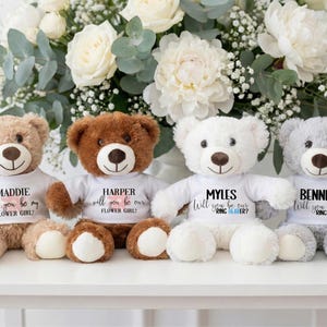 May include: Four plush teddy bears in various colors: tan, brown, white, and gray. Each bear wears a white t-shirt with a personalized message. The text on the shirts reads "MADDIE will you be my FLOWER GIRL?", "HARPER will you be our FLOWER GIRL?", "MYLES Will you be our RING BEARER?", and "BENNETT Will you be our RING BEARER?"