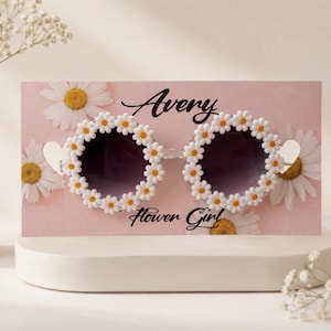 May include: Daisy-shaped sunglasses with dark lenses and white daisy accents. The sunglasses are connected by a small heart-shaped detail. The text "Avery Flower Girl" is written on a pink background with daisy accents.