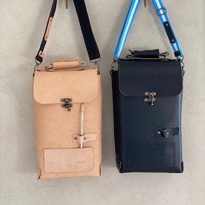 K&ouml;nnte beinhalten: Zwei Lederrucks&auml;cke h&auml;ngen an Haken. Der Rucksack links ist aus braunem Leder mit einem schwarzen Riemen und einer kleinen Tasche vorne. Der Rucksack rechts ist aus schwarzem Leder mit einem blau-wei&szlig; gestreiften Riemen und einer kleinen Tasche vorne. Beide Rucks&auml;cke haben einen Verschluss mit einer Messing-Schnalle.