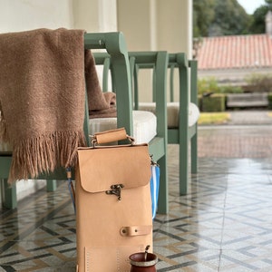K&ouml;nnte beinhalten: Eine hellbraune Ledertasche mit einem Messingverschluss und einem blau-wei&szlig; gestreiften Trageriemen. Die Tasche steht auf einem gefliesten Boden mit einer braunen Holz-Mate-Tasse und einer Bombilla.