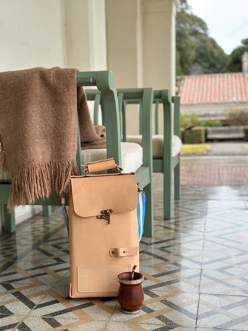 K&ouml;nnte beinhalten: Eine hellbraune Ledertasche mit einem Messingverschluss und einem blau-wei&szlig; gestreiften Trageriemen. Die Tasche steht auf einem gefliesten Boden mit einer braunen Holz-Mate-Tasse und einer Bombilla.