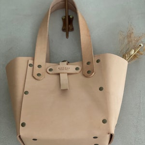 K&ouml;nnte beinhalten: Eine hellbraune Ledertasche mit einem Schnallenverschluss und Metallnieten. Die Tasche hat zwei Henkel und der Markenname "ANNONNI" ist auf einem Lederanh&auml;nger aufgedruckt.