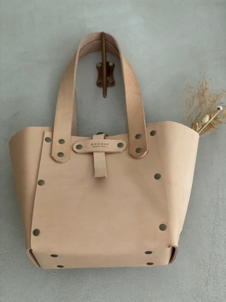 K&ouml;nnte beinhalten: Eine hellbraune Ledertasche mit einem Schnallenverschluss und Metallnieten. Die Tasche hat zwei Henkel und der Markenname "ANNONNI" ist auf einem Lederanh&auml;nger aufgedruckt.