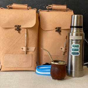 K&ouml;nnte beinhalten: Zwei braune Lederrucks&auml;cke mit einem Metallverschluss und einer kleinen Tasche auf der Vorderseite. Eine silberne Thermoskanne mit einem Aufkleber, auf dem "Van Cal Ven" steht, befindet sich neben den Rucks&auml;cken. Ein brauner Holzbecher mit silbernem Rand und einem silbernen Strohhalm steht vor der Thermoskanne. Ein blaues und wei&szlig; gestreiftes Band liegt vor den Rucks&auml;cken.