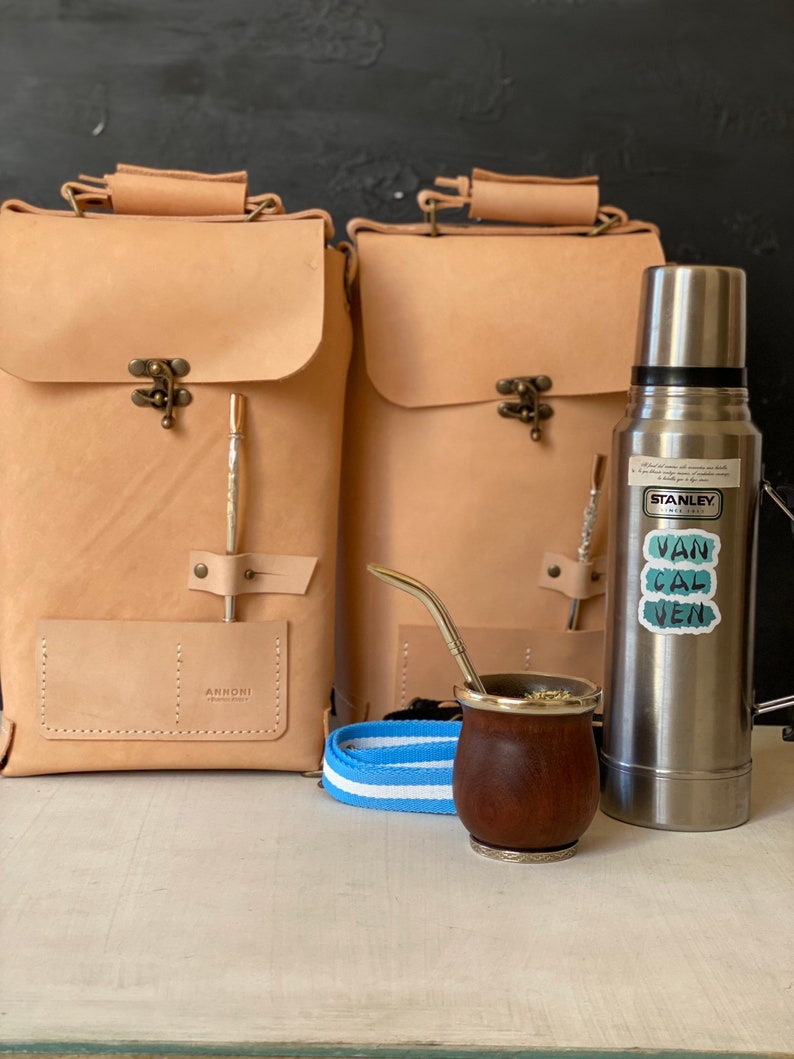 K&ouml;nnte beinhalten: Zwei braune Lederrucks&auml;cke mit einem Metallverschluss und einer kleinen Tasche auf der Vorderseite. Eine silberne Thermoskanne mit einem Aufkleber, auf dem "Van Cal Ven" steht, befindet sich neben den Rucks&auml;cken. Ein brauner Holzbecher mit silbernem Rand und einem silbernen Strohhalm steht vor der Thermoskanne. Ein blaues und wei&szlig; gestreiftes Band liegt vor den Rucks&auml;cken.