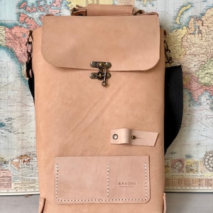 K&ouml;nnte beinhalten: Eine hellbraune Ledertasche mit einem Klappverschluss und einer kleinen Fronttasche. Die Tasche hat einen schwarzen verstellbaren Riemen und eine Messing-Schnalle. Die Tasche ist mit "ANNONNI Buenos Aires" beschriftet.