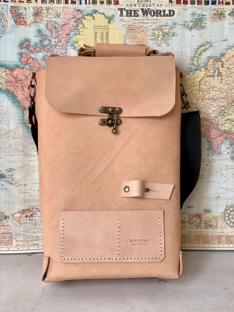 K&ouml;nnte beinhalten: Eine hellbraune Ledertasche mit einem Klappverschluss und einer kleinen Fronttasche. Die Tasche hat einen schwarzen verstellbaren Riemen und eine Messing-Schnalle. Die Tasche ist mit "ANNONNI Buenos Aires" beschriftet.