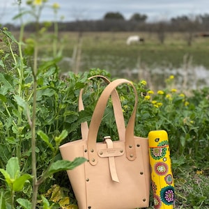 K&ouml;nnte beinhalten: Eine hellbraune Ledertasche mit einer Schlie&szlig;e und einer gelben Thermoskanne mit Blumenmuster. Die Tasche hat ein Etikett mit der Aufschrift "Armoni" und steht auf einem gr&uuml;nen Grasbett.