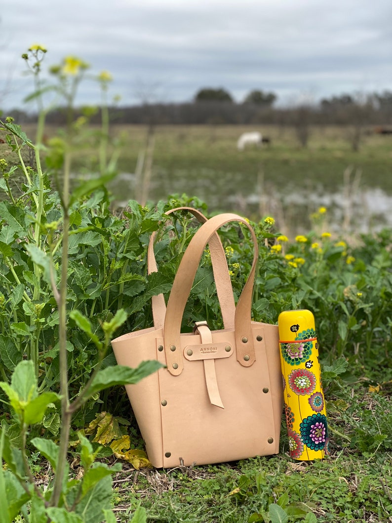 K&ouml;nnte beinhalten: Eine hellbraune Ledertasche mit einer Schlie&szlig;e und einer gelben Thermoskanne mit Blumenmuster. Die Tasche hat ein Etikett mit der Aufschrift "Armoni" und steht auf einem gr&uuml;nen Grasbett.