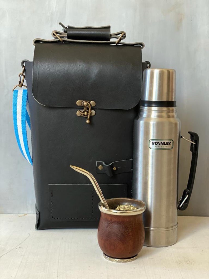 K&ouml;nnte beinhalten: Eine schwarze Ledertasche mit einem blau-wei&szlig; gestreiften Trageriemen, eine silberne Thermoskanne mit dem Text "STANLEY SINCE 1913" und eine Mate-Trinktasse aus Holz mit einem Metallstrohhalm.