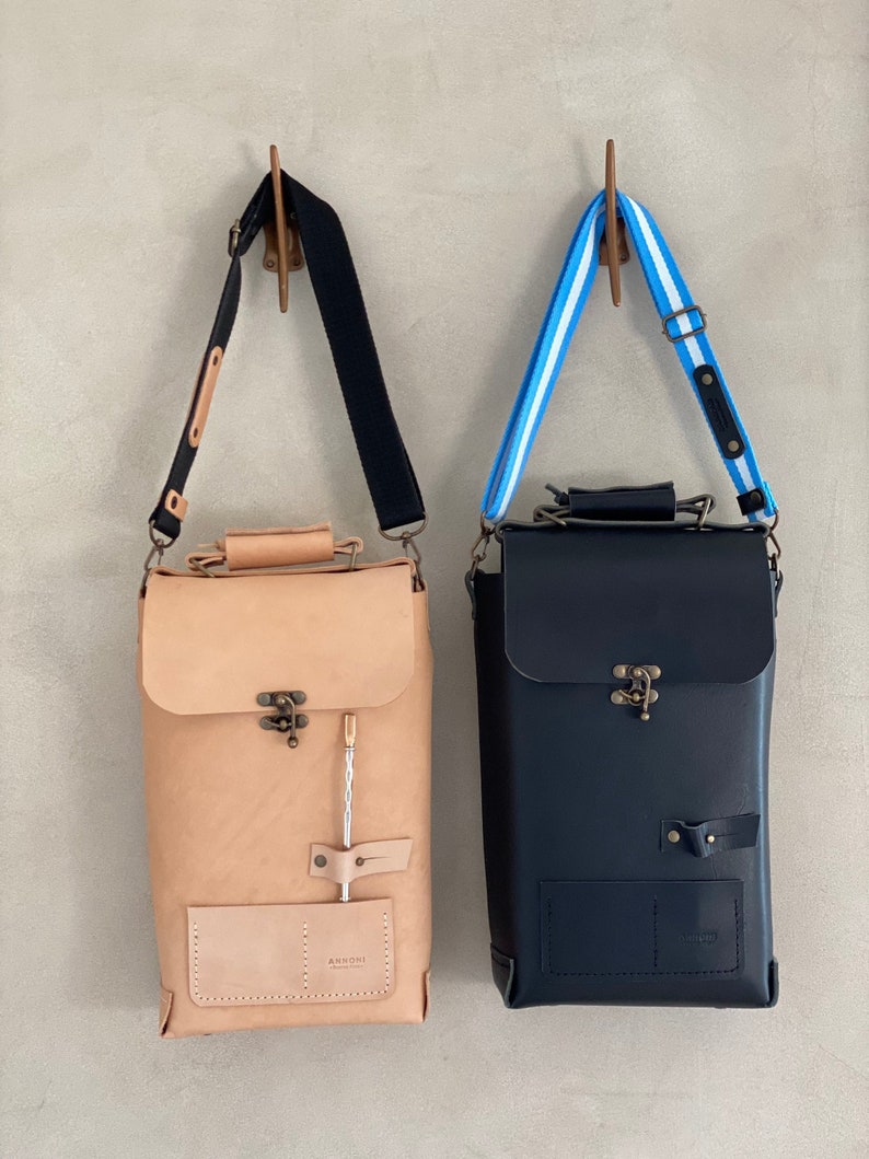 K&ouml;nnte beinhalten: Zwei Lederrucks&auml;cke h&auml;ngen an Haken. Der Rucksack links ist aus braunem Leder mit einem schwarzen Riemen und einer kleinen Tasche vorne. Der Rucksack rechts ist aus schwarzem Leder mit einem blau-wei&szlig; gestreiften Riemen und einer kleinen Tasche vorne. Beide Rucks&auml;cke haben einen Verschluss mit einer Messing-Schnalle.