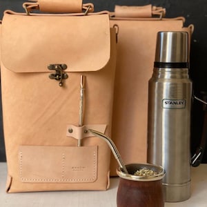 K&ouml;nnte beinhalten: Ein Set aus zwei braunen Ledertaschen mit einem Metallverschluss, einer silbernen Thermoskanne mit dem Schriftzug "STANLEY", einer Holztasse mit silbernem Rand und einem Metallstrohhalm.