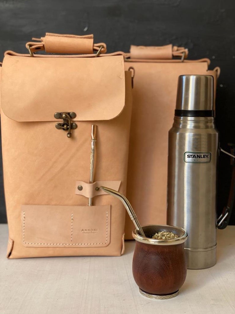 K&ouml;nnte beinhalten: Ein Set aus zwei braunen Ledertaschen mit einem Metallverschluss, einer silbernen Thermoskanne mit dem Schriftzug "STANLEY", einer Holztasse mit silbernem Rand und einem Metallstrohhalm.