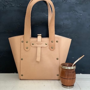 K&ouml;nnte beinhalten: Eine hellbraune Ledertasche mit einem Riemen und Metallnieten. Die Tasche tr&auml;gt den Text "ANNONII" auf einem Metallschild. Im Vordergrund steht ein Holzbecher mit einem silbernen Band und einem Strohhalm.