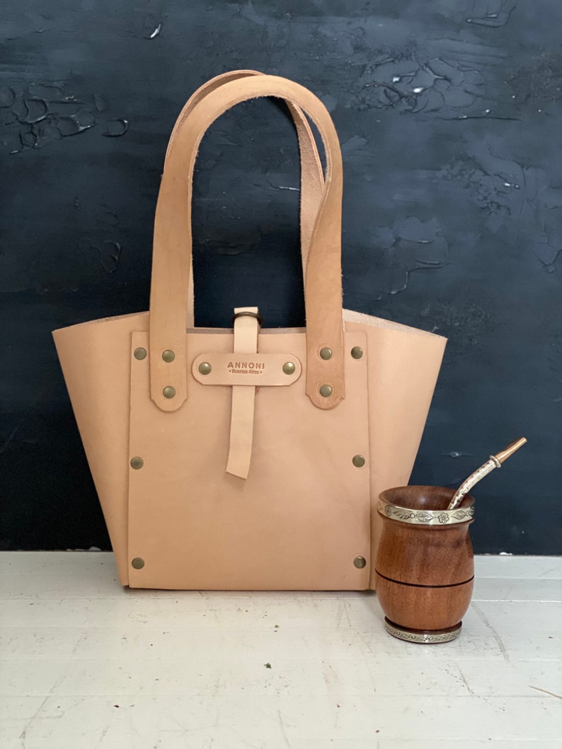 K&ouml;nnte beinhalten: Eine hellbraune Ledertasche mit einem Riemen und Metallnieten. Die Tasche tr&auml;gt den Text "ANNONII" auf einem Metallschild. Im Vordergrund steht ein Holzbecher mit einem silbernen Band und einem Strohhalm.