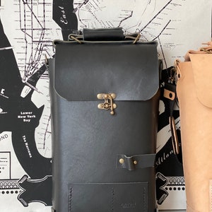 K&ouml;nnte beinhalten: Eine schwarze Ledertasche mit einem Messingverschluss und einem kleinen Fach auf der Vorderseite. Die Tasche hat einen Riemen und wird von Airmail Products hergestellt.