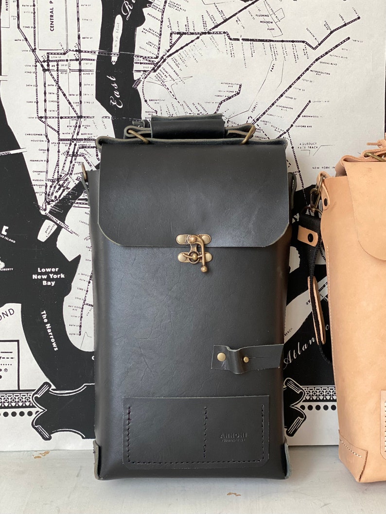 K&ouml;nnte beinhalten: Eine schwarze Ledertasche mit einem Messingverschluss und einem kleinen Fach auf der Vorderseite. Die Tasche hat einen Riemen und wird von Airmail Products hergestellt.