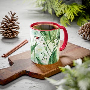 May include: A red and white coffee mug with a floral design featuring lily of the valley flowers and a ladybug. The mug is filled with coffee and is sitting on a wooden cutting board.