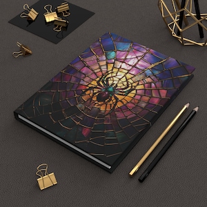 May include: A black notebook with a colorful stained glass spider web design. The spider web is made of purple, blue, green, and gold glass pieces. A black spider is in the center of the web.