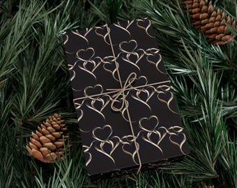 May include: A wrapped gift with a black background and a pattern of gold hearts. The gift is tied with twine and rests on a bed of green pine needles, with pine cones scattered around.