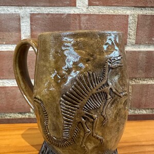 May include: A brown ceramic mug featuring a raised dinosaur skeleton design. The mug has a handle and sits on a decorative base. The dinosaur skeleton is detailed, adding texture to the mug's surface. The mug is suitable for hot beverages.