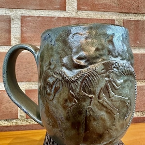 Peut inclure: Une tasse en céramique artisanale avec un motif de squelette de dinosaure. La tasse a un glaçage texturé vert et bleu terreux et une grande anse. La tasse mesure environ 10 cm de haut et 13 cm de large.