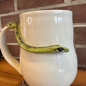 May include: Cream ceramic mug featuring a sculpted green and yellow snake wrapped around the body. The snake's head faces the viewer. The mug has a rounded base and a large handle.