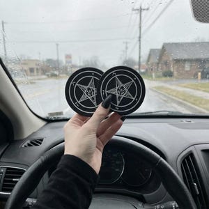 May include: Two black round car coasters with a white pentagram and goat head design. The coasters are held up in front of a car's windshield on a rainy day.