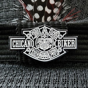 May include: A black and white enamel pin with the words "Chicano Biker" and a stylized Aztec design in the center.