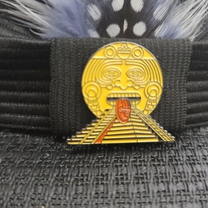 May include: A gold-colored metal pin with a depiction of a Mayan pyramid and a face with a red tongue. The pin is on a black fabric background.