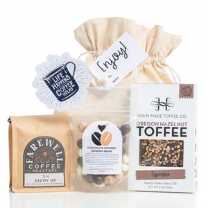 May include: A gift set featuring coffee, toffee, and chocolate-covered espresso beans. The set includes a bag of coffee, a box of Oregon Hazelnut Toffee, and a clear bag of chocolate-covered espresso beans, with a tag that says "Enjoy!"