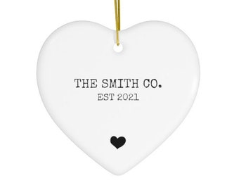 May include: A white heart-shaped ornament with the text "THE SMITH CO. EST 2021" and a black heart graphic. A gold string is attached for hanging. The ornament is likely ceramic or porcelain.