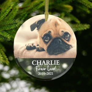 May include: Round glass Christmas ornament featuring a pug dog. The ornament has a gold ribbon for hanging. The text "CHARLIE Forever Loved 2010-2021" is printed on the bottom of the ornament.