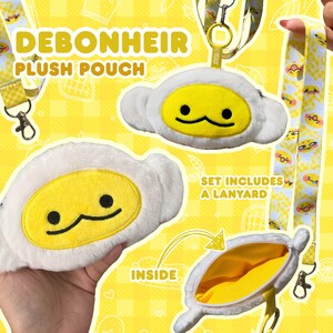 Può includere: Un set di pochette in peluche con un design a uovo giallo e bianco. Il set include un cordino con un motivo abbinato. La pochette ha una chiusura a cerniera e il testo "DEBONHEIR PLUSH POUCH" è visibile.