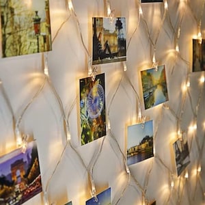 May include: A string of white fairy lights with small, warm white bulbs. The lights are strung across a white wall and have photos clipped to them with small clothespins. The photos are of various sizes and subjects, including a city scene, a person sitting, and a Ferris wheel.